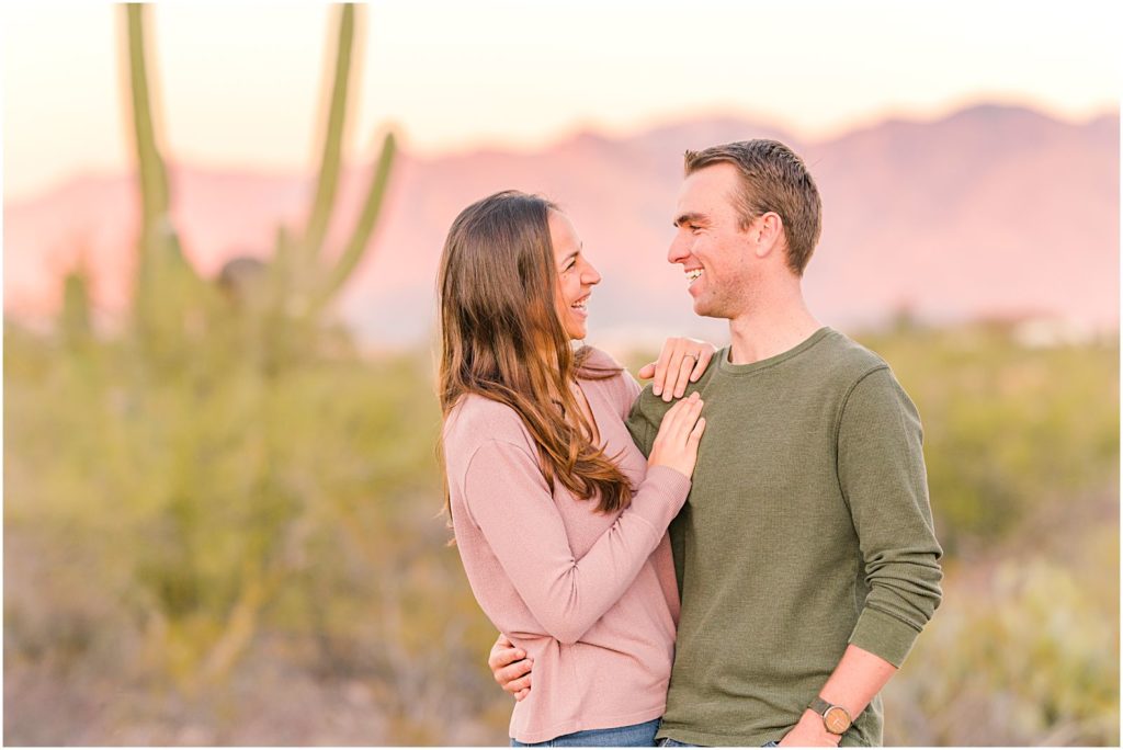engaged couple smiling at each other with pink mountains behind them at sunset