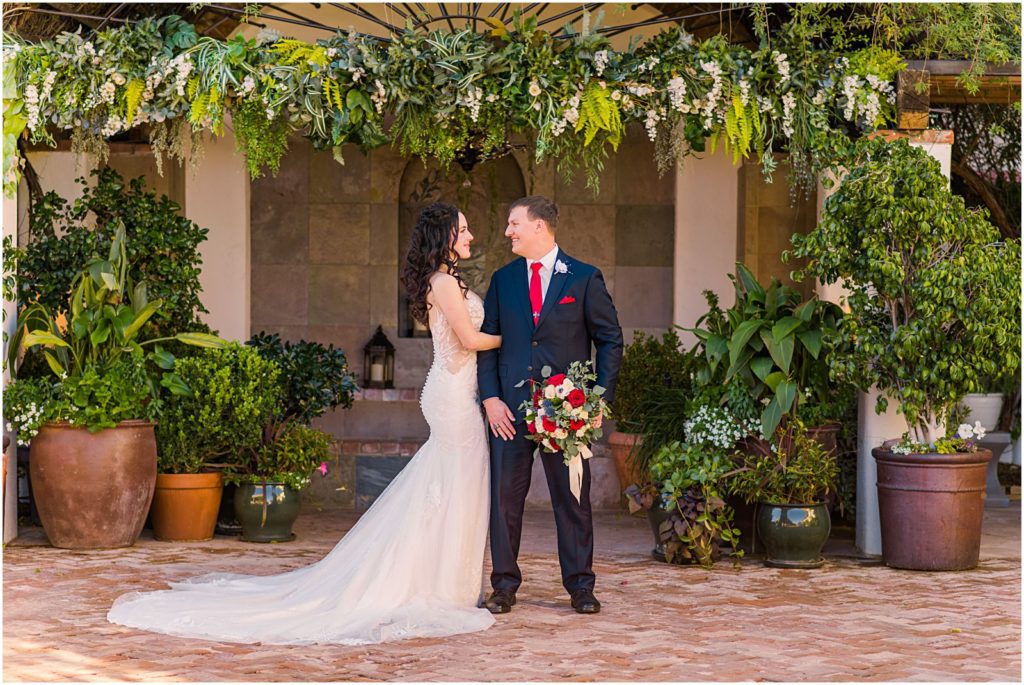pulled back portrait of bride and groom under arbor covered in greenery in courtyard