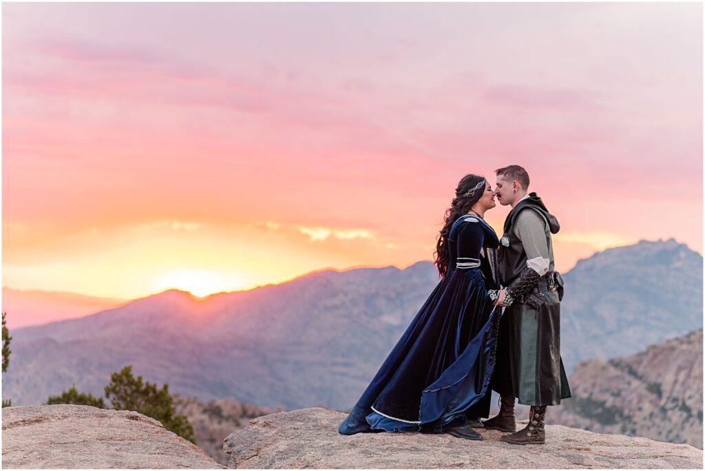 bride and groom kiss on mountaintop with colorful sunset at fantasy themed wedding